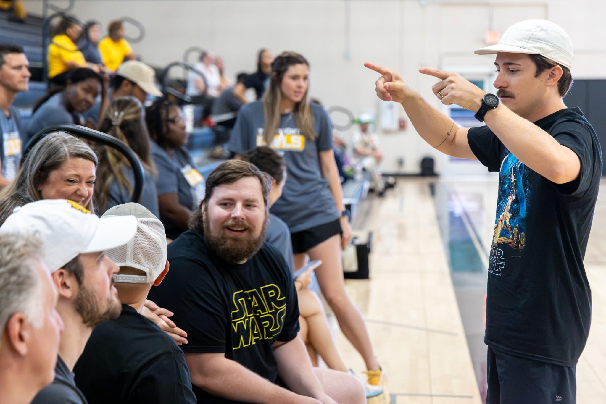 2026 Owlympics / At a lively indoor event, a mustached man in a white cap speaks and gestures to seated attendees, including a bearded Star Wars fan.