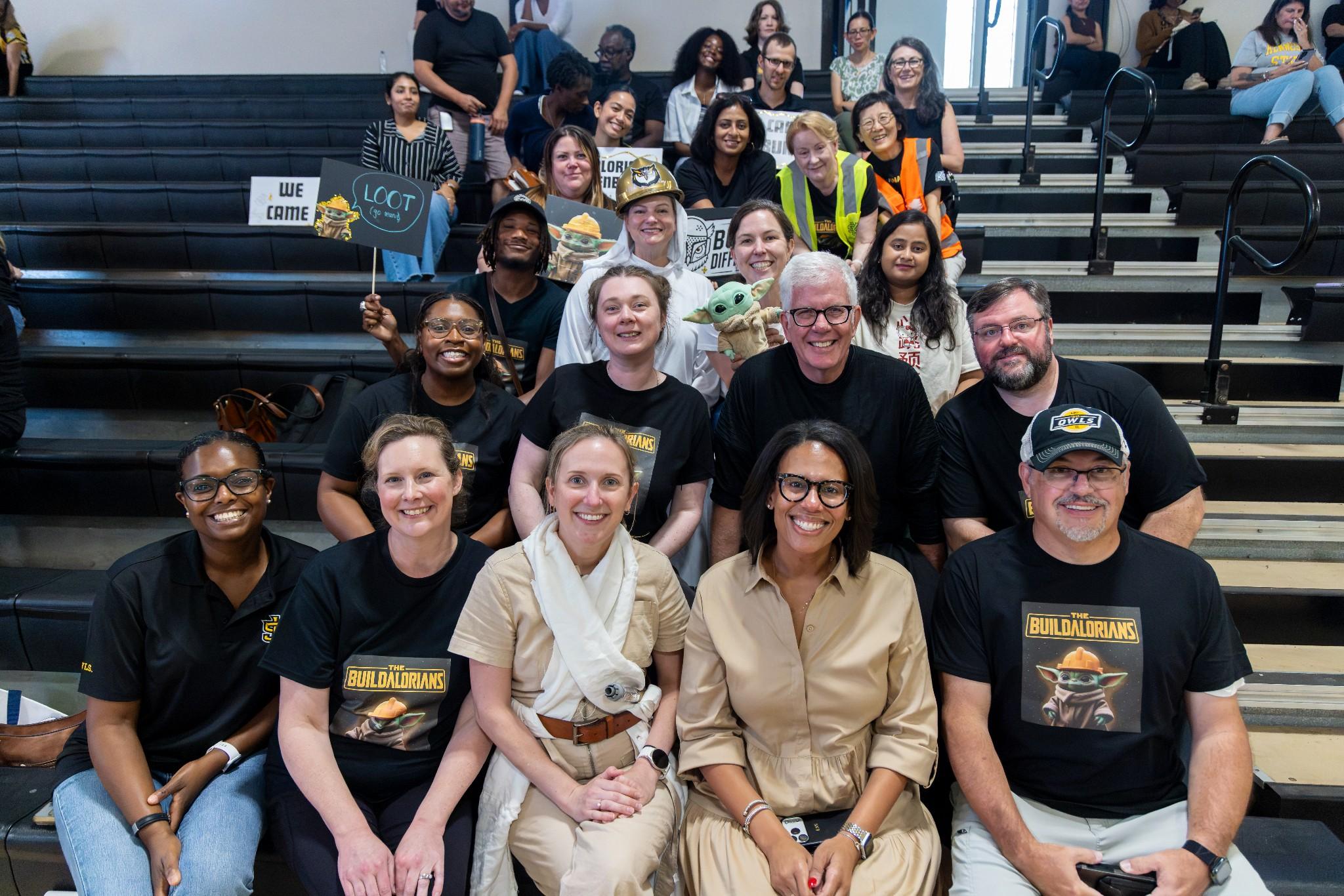 2026 Owlympics / A diverse, smiling group poses on gymnasium bleachers, cheering on the Owlympians and holding signs, creating a warm, celebratory team atmosphere.