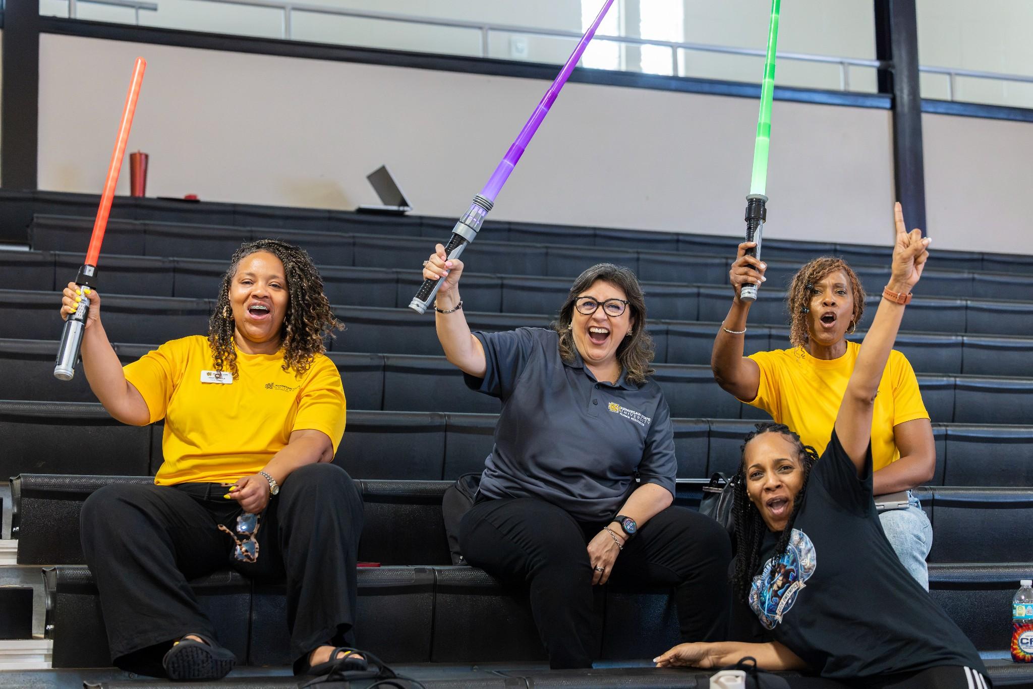 2026 Owlympics / Four enthusiastic women sit in gymnasium bleachers, cheering and raising colorful light-up wands, creating a joyful, energetic mood.