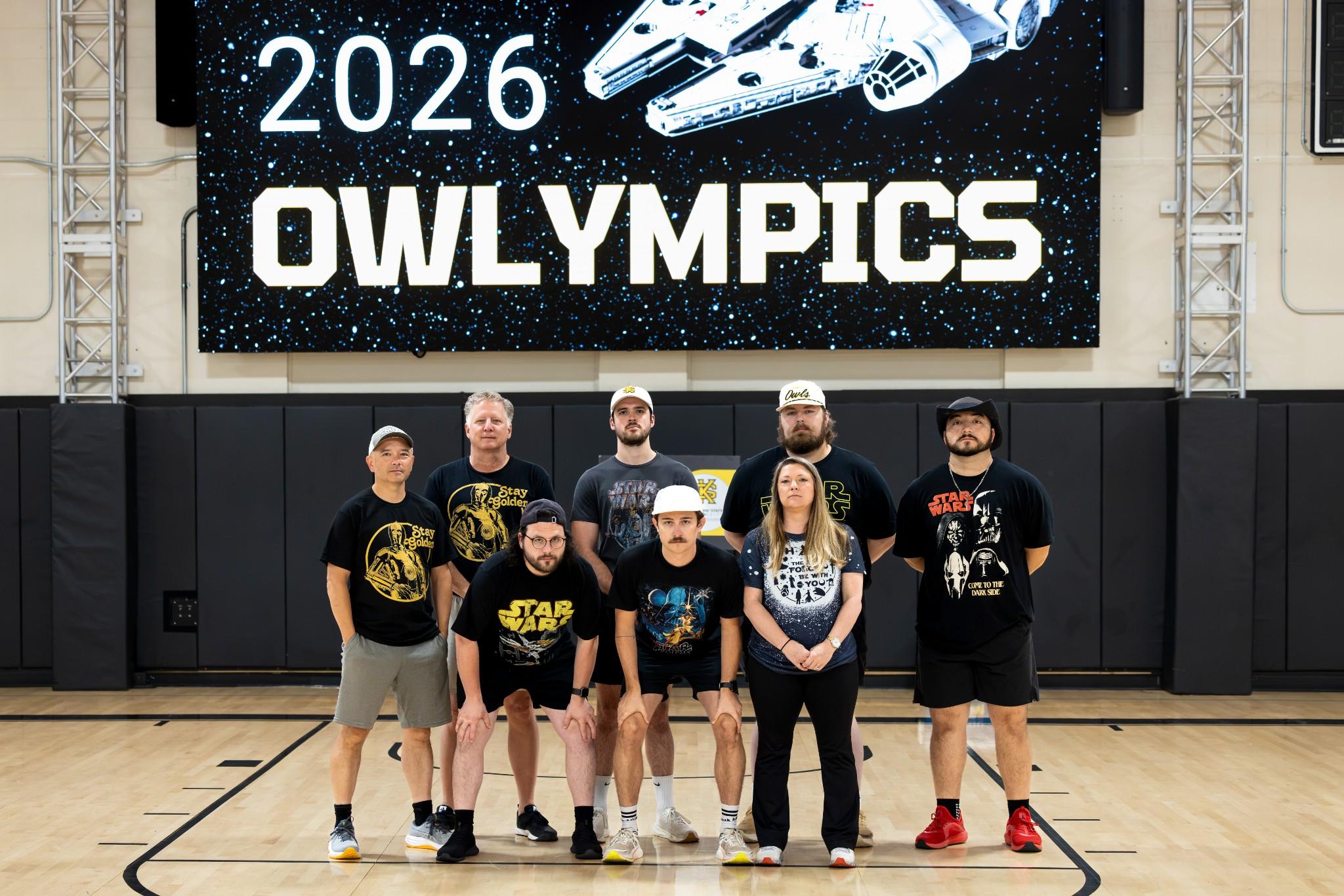 2026 Owlympics / Seven people pose on a basketball court beneath a space-themed “2026 OWLympics” banner, creating a fun, energetic team atmosphere