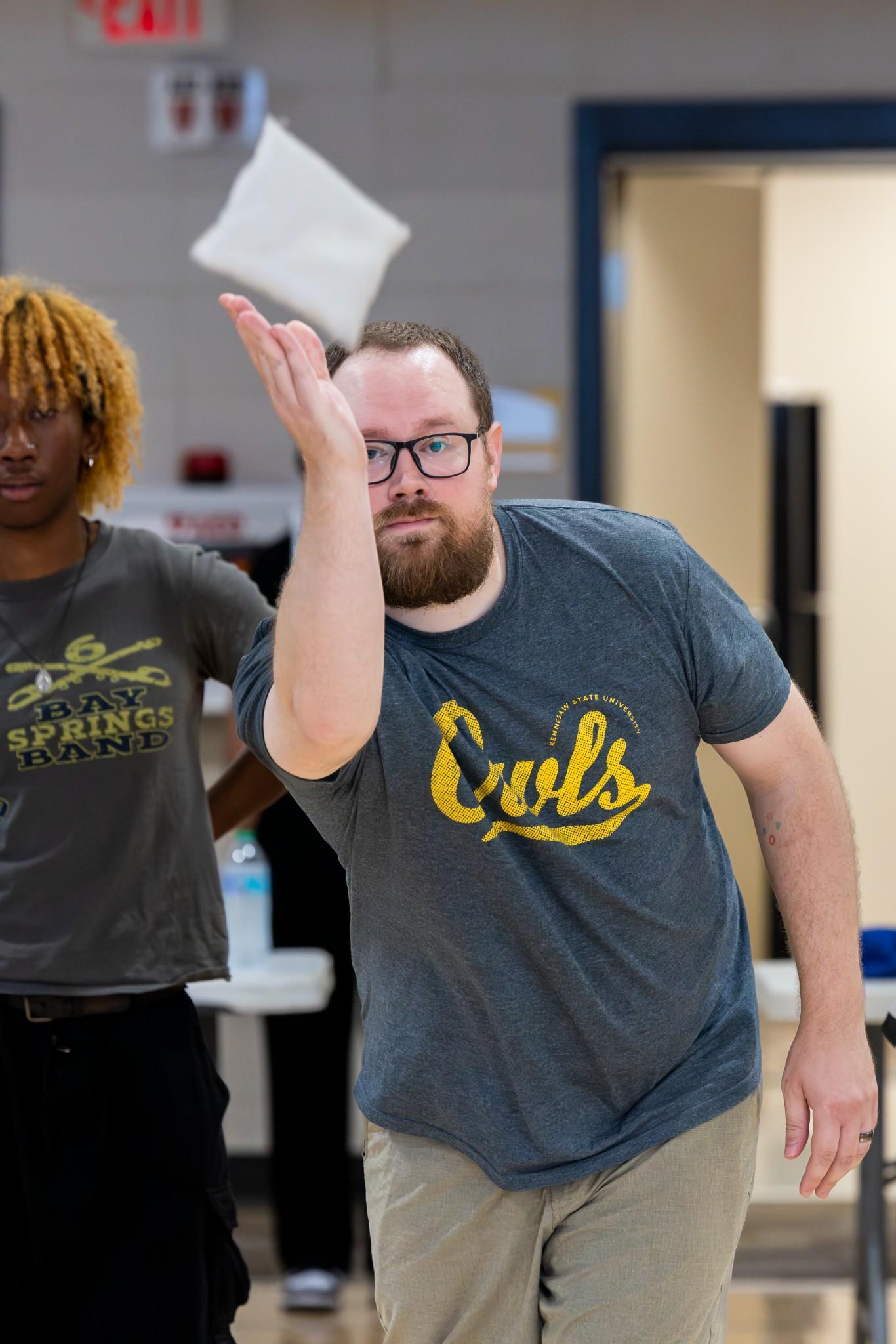 2026 Owlympics / Bearded man in glasses tosses bean bag during owlympics game, watched by a person with dreadlocks; playful, focused mood.