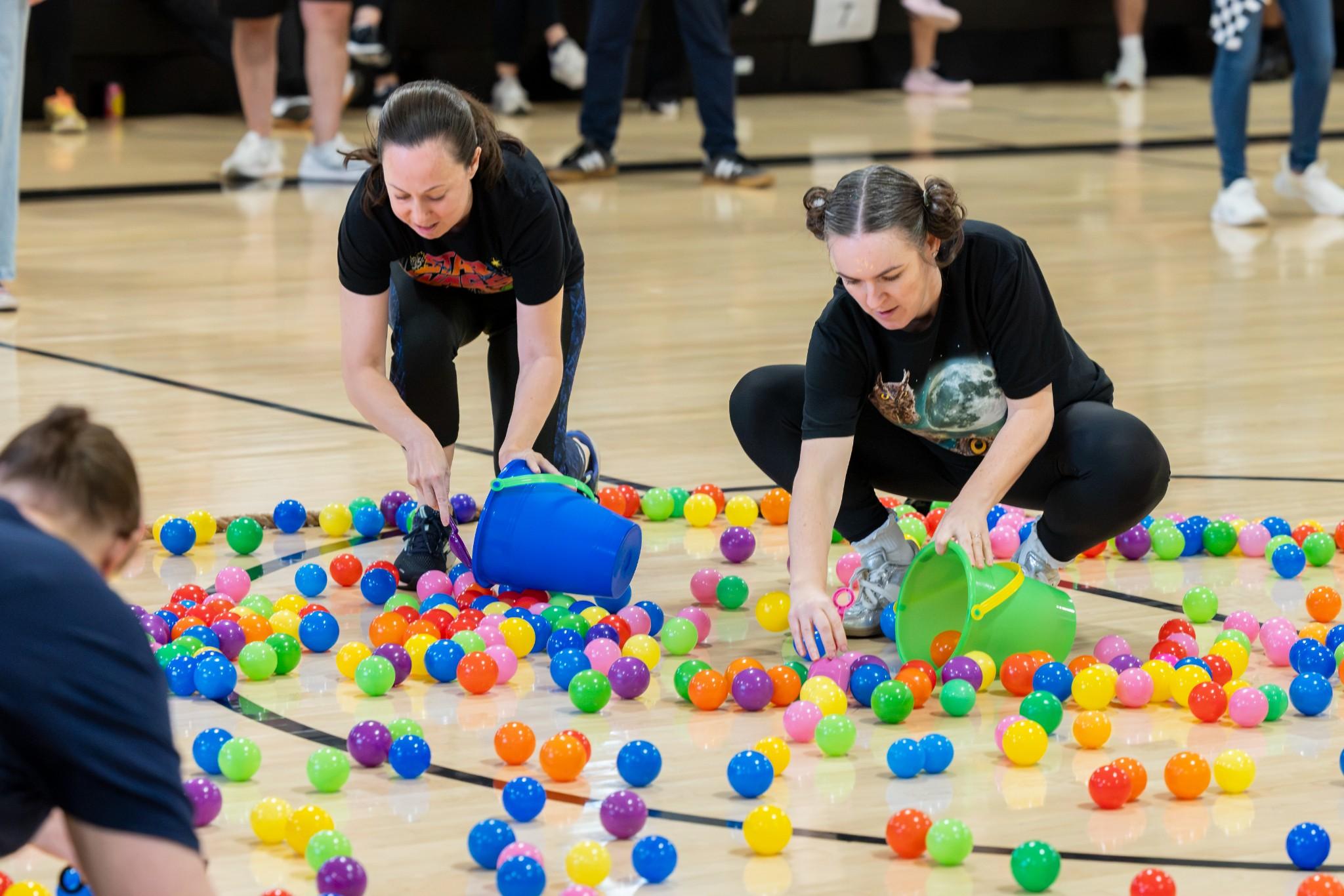 2026 Owlympics / Two women in a gym gather colorful plastic balls from the floor, creating a lively, playful scene during a group activity.