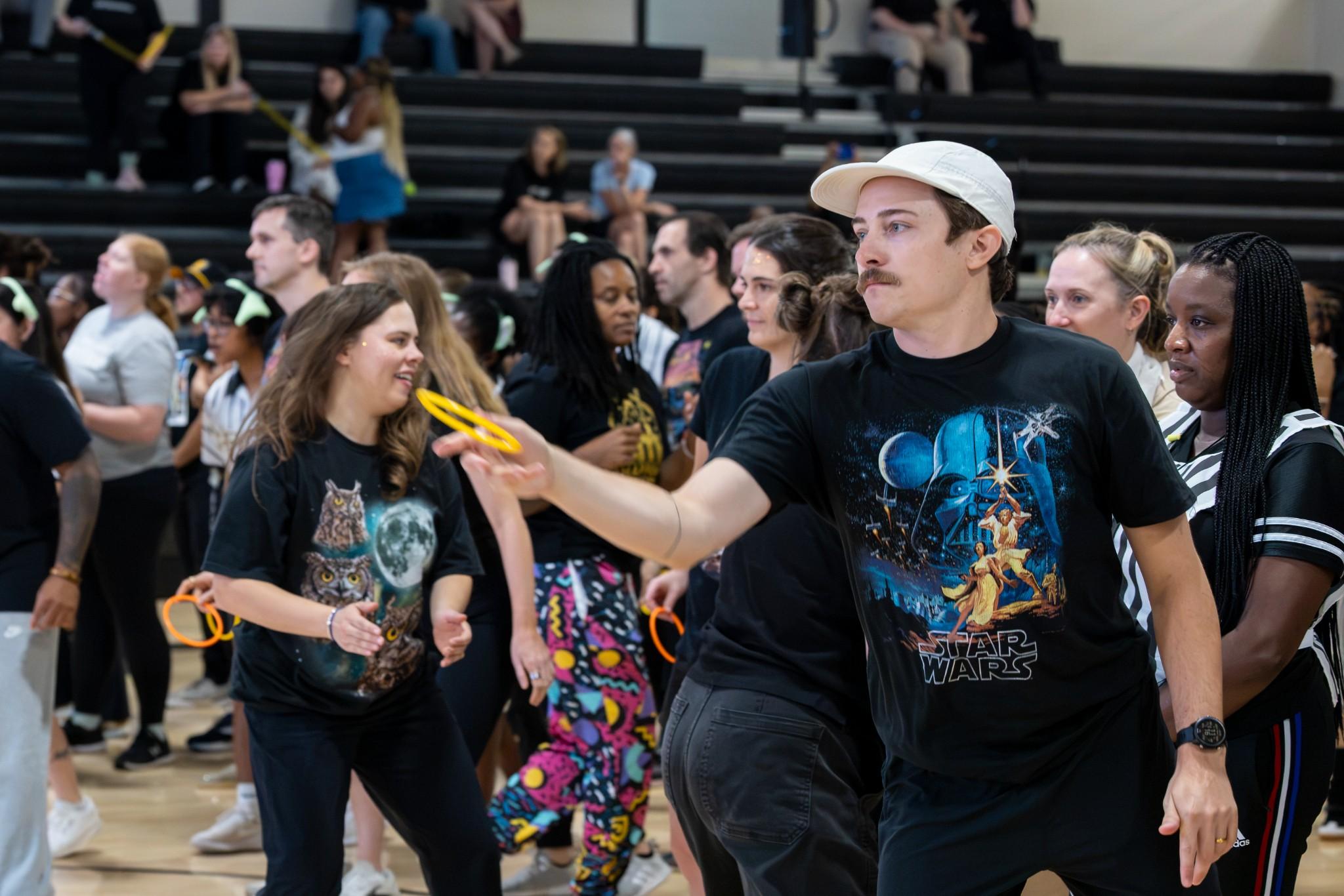 2026 Owlympics / Group of people in a gymnasium; a man in a white cap holds yellow glow sticks, creating a lively, joyful atmosphere.