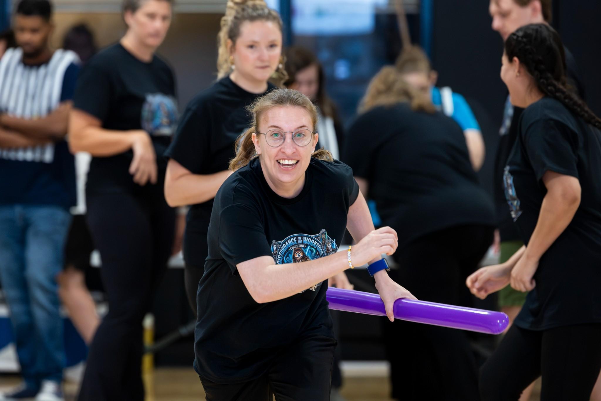 2026 Owlympics / Smiling woman in black sportswear holds a purple light saber during a lively group activity, surrounded by participants in a gym.