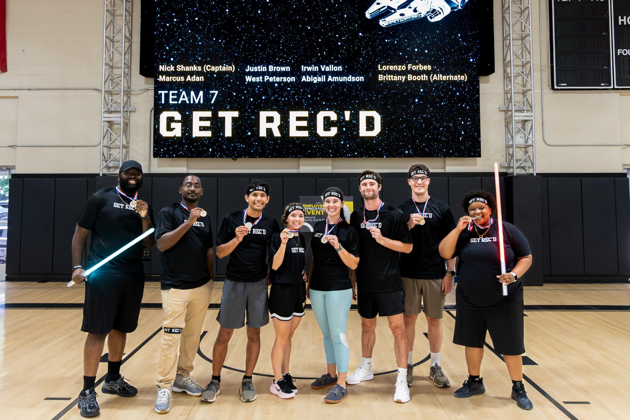 2026 Owlympics / Seven smiling teammates pose on a basketball court beneath a screen reading “Team 7: Get Rec’d,” celebrating with playful lightsabers.
