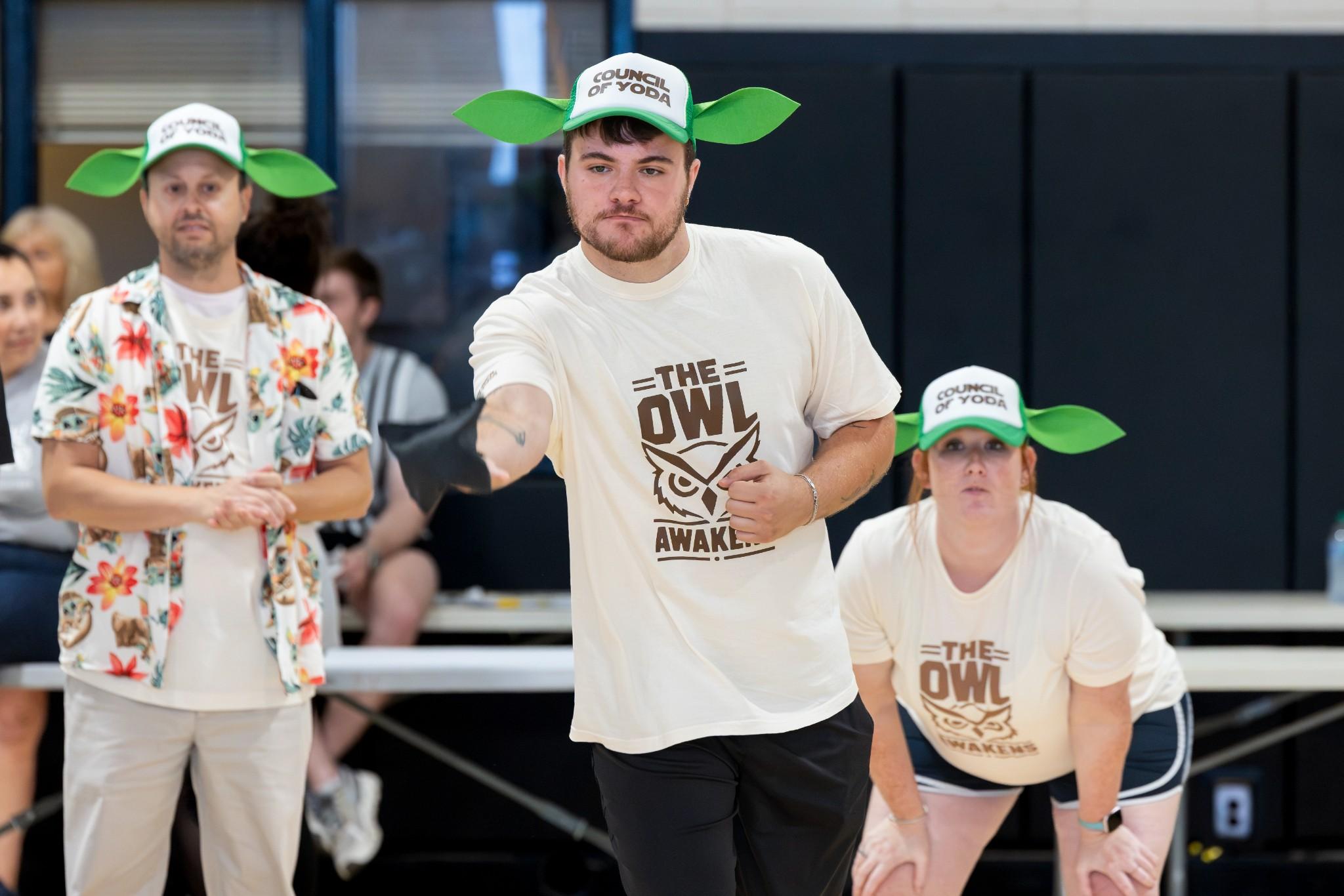 2026 Owlympics / Three team members in owl-themed shirts and grogu caps watch intently while the center man stepping forward to toss a bean bag.