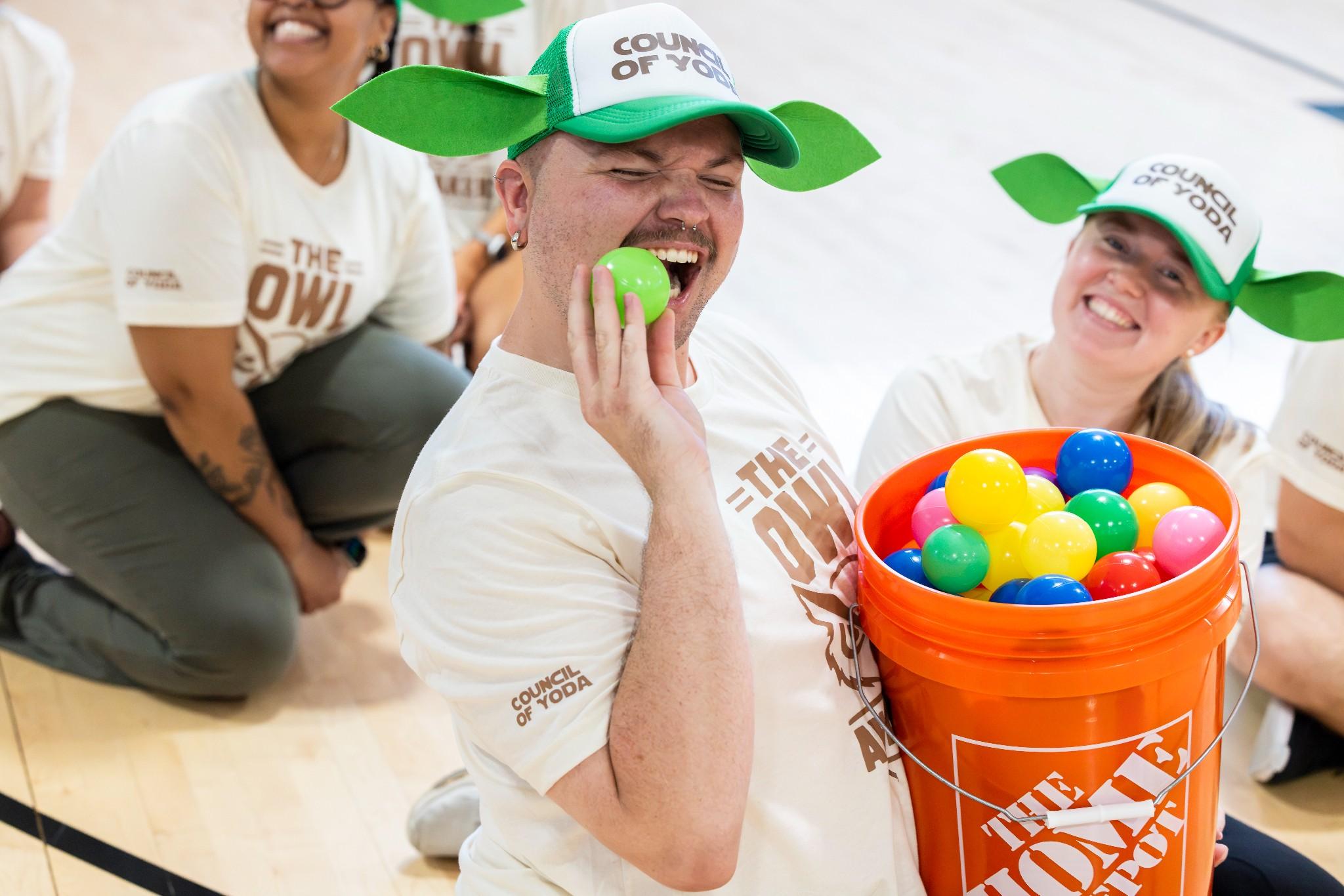 2026 Owlympics / Joyful volunteers in grogu hats laugh during Owlympics, as one pretends to bite a green ball beside a bucket of colorful balls.