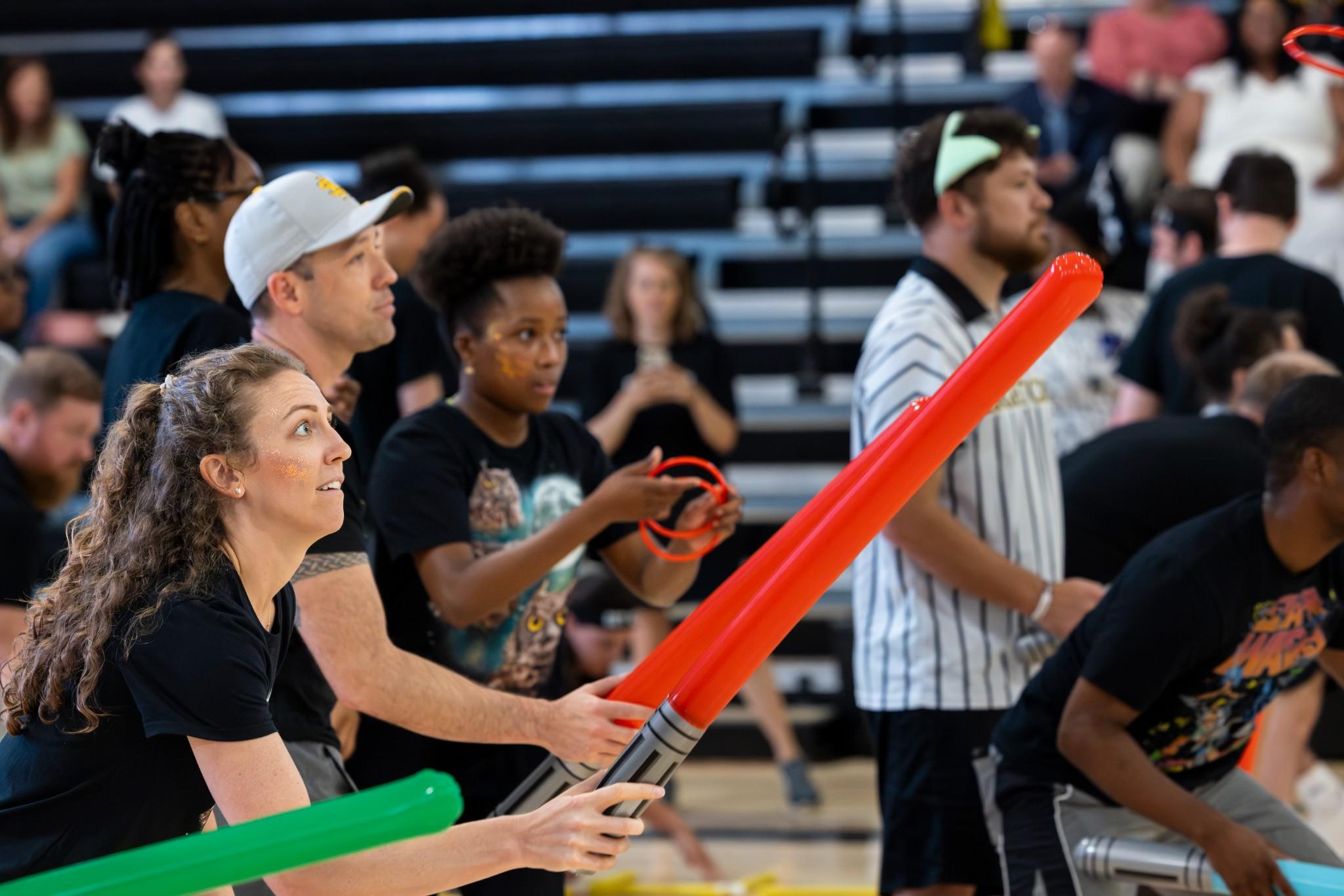 2026 Owlympics / Players in a lively gym use colorful light sabers during an indoor team game; a woman reaches forward while spectators watch from bleachers.