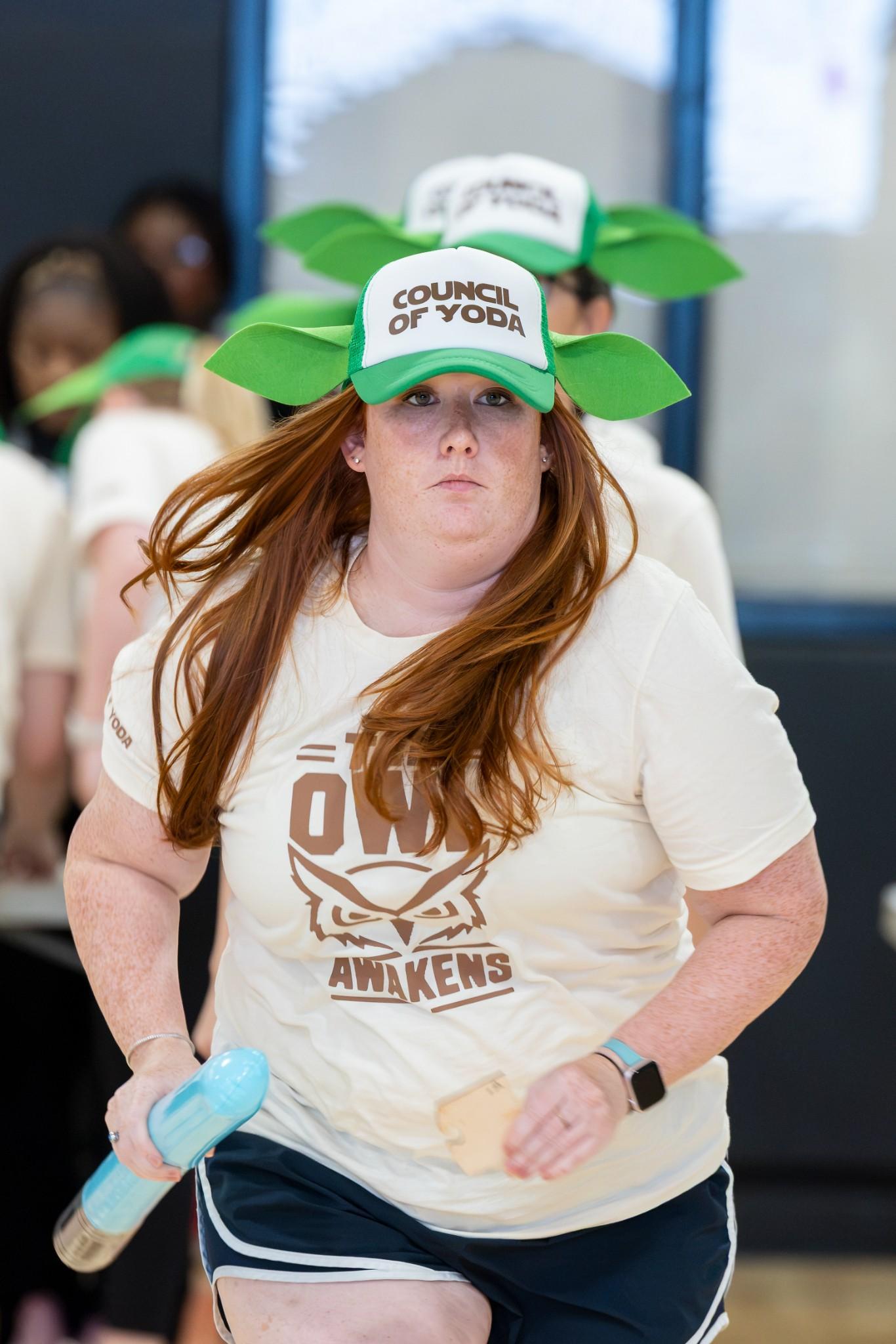 2026 Owlympics / Red-haired woman runs indoors wearing a green “Council of Yoda” hat and Star Wars shirt, gripping a blue light saber with an energetic expression.
