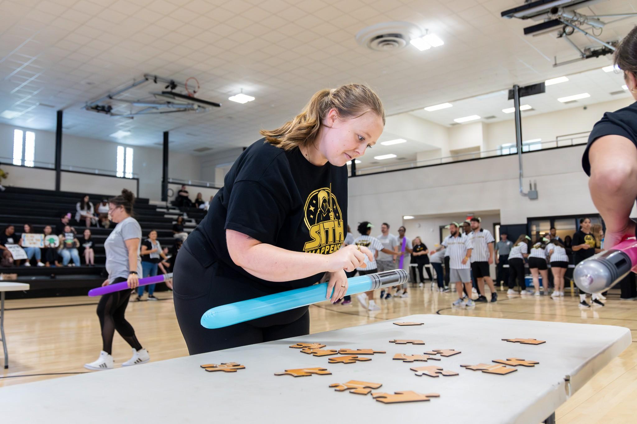 2026 Owlympics / In a busy school gym, a smiling girl in a black shirt holds a blue light saber while working on a wooden puzzle pieces at a community activity table.