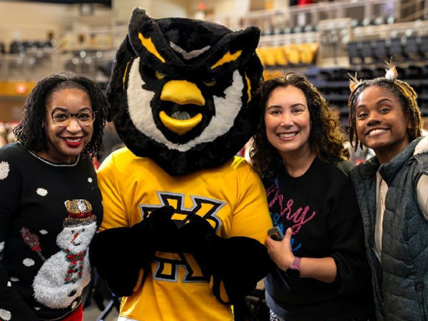 Four people smiling, including a person in an owl mascot costume wearing a yellow shirt, are gathered in a stadium setting, exuding a joyful atmosphere.