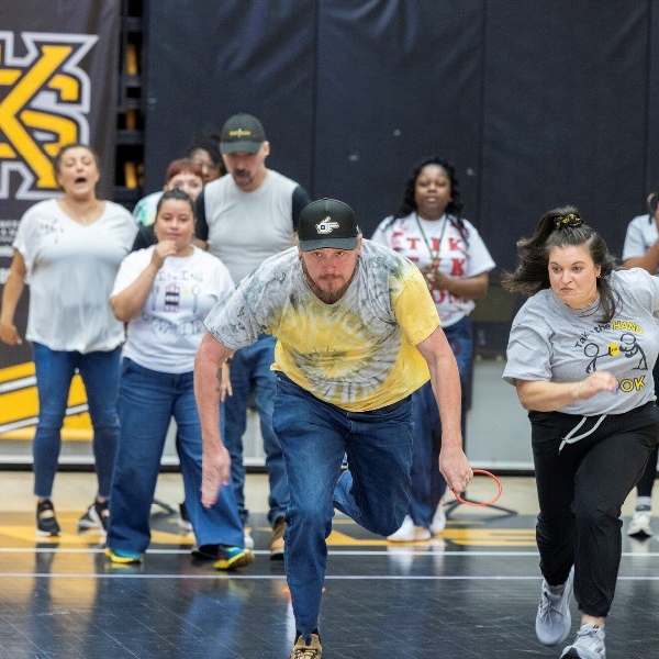 People in casual clothing race energetically on an indoor court, some cheering. Background features a "KS" banner, adding a competitive tone.