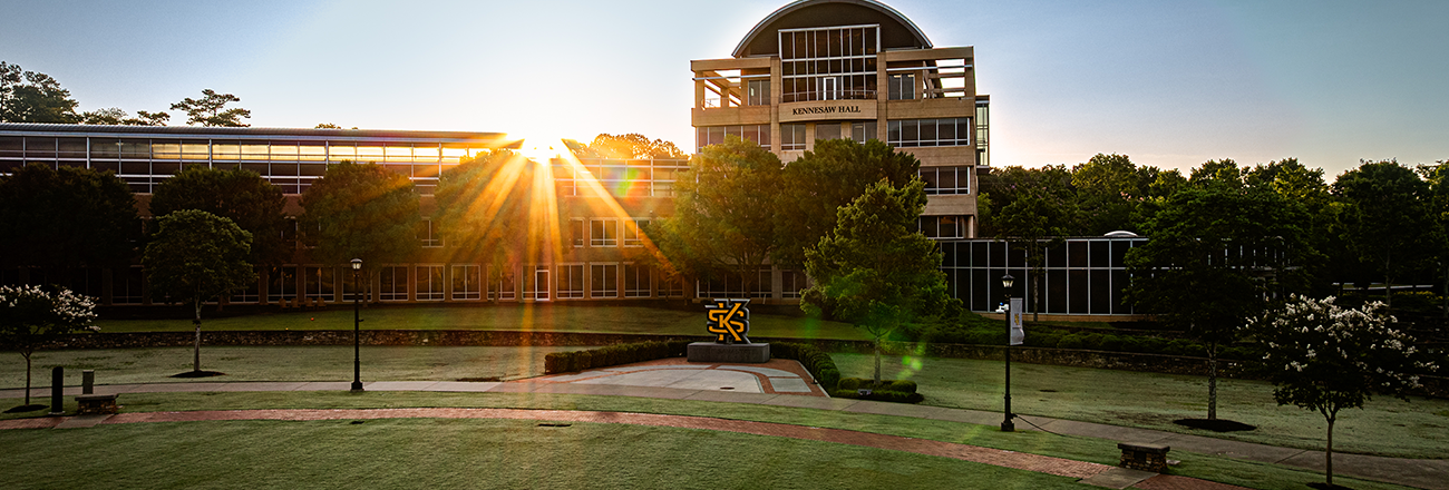 Sunrise over kennesaw hall with a glass facade, surrounded by trees. Warm light casts long shadows, creating a serene and hopeful ambiance.