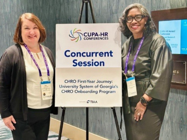 Two women stand smiling beside a sign for a CUPA-HR conference session on CHRO onboarding. Both wear conference badges and purple lanyards.