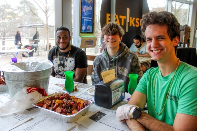 three male students eating crawfish in the dining hall and smiling