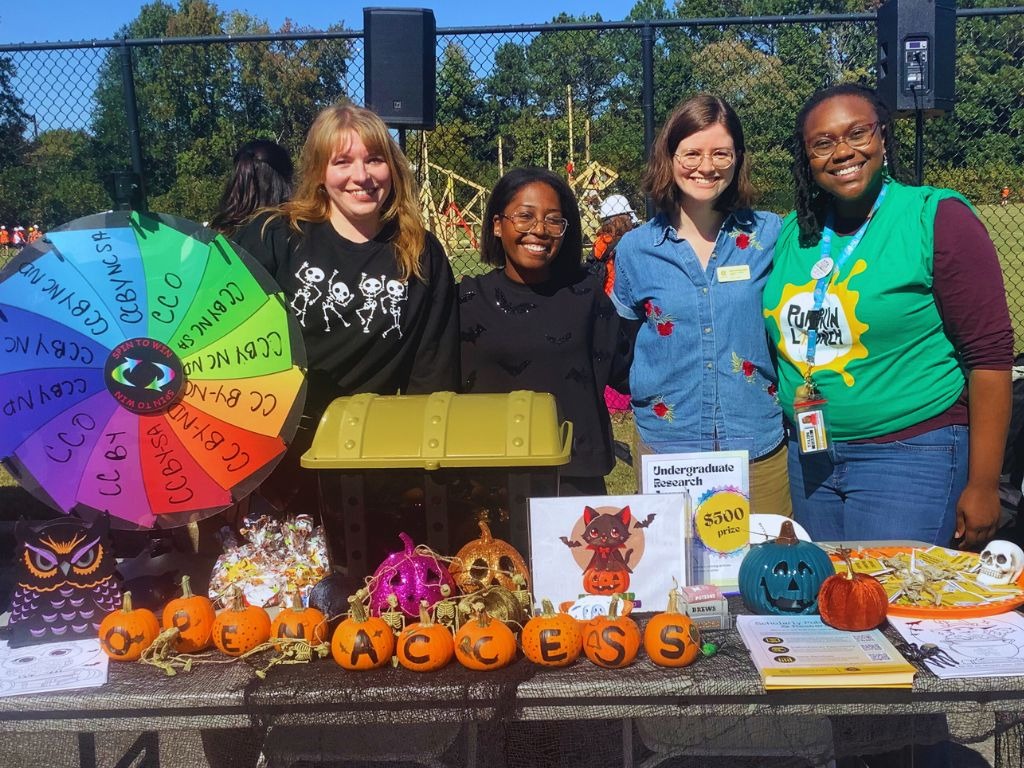 Four people standing behind a Halloween themed event table with a tresure chest, a multi-colorednprize wheel, and a variety of brochures.