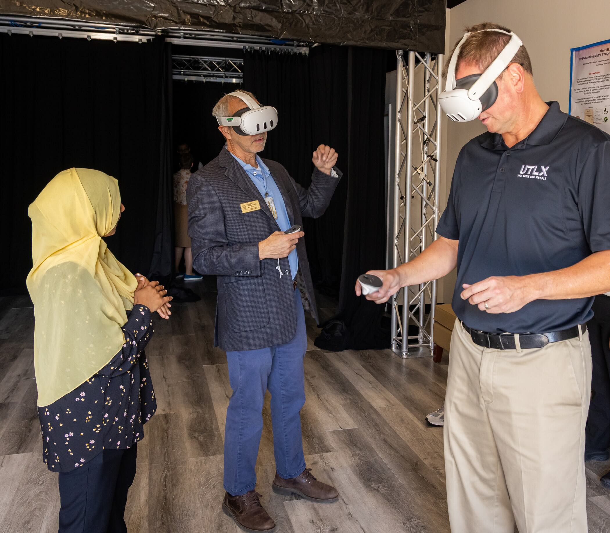 The image captures a moment in a virtual reality (VR) demonstration setting. Three individuals are present in a spacious area, equipped with VR headsets.