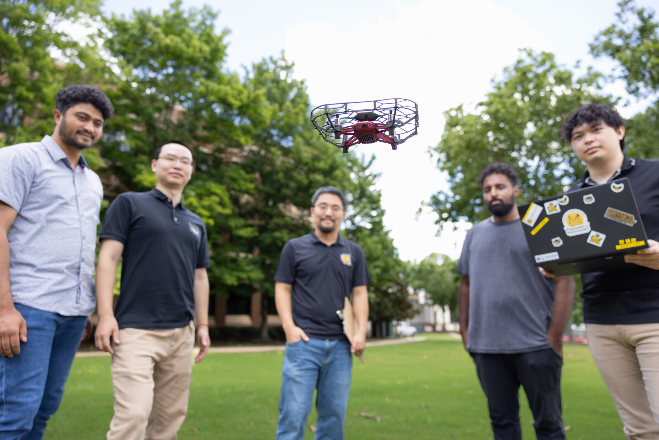 Five people stand on a grassy area outdoors, watching a small drone flying in the center. One person holds a laptop decorated with stickers. Trees and buildings are visible in the background.