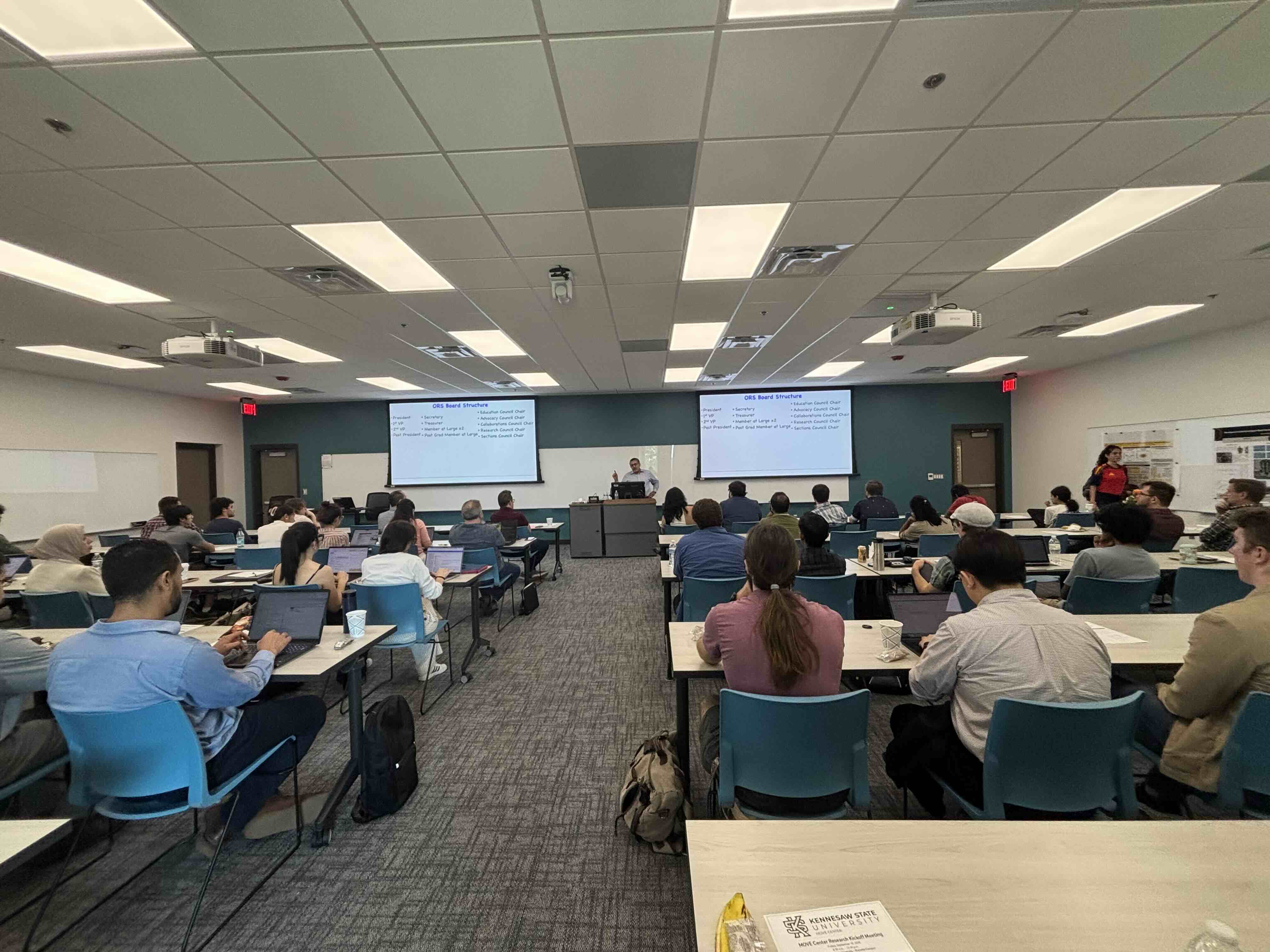 A presenter addresses a seated audience in a classroom while two screens display an “ORS Board Structure” slide.