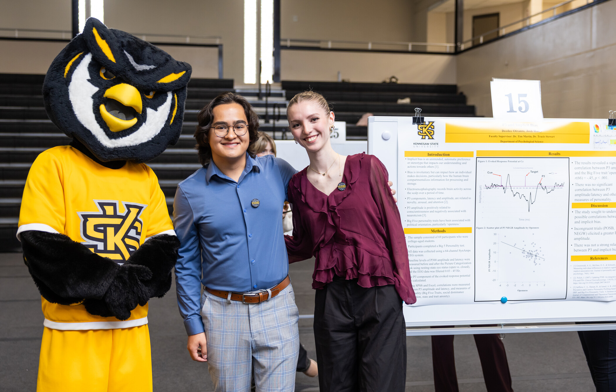 Three people, including a person in a Kennesaw State University owl mascot suit, stand by a research poster in a gymnasium, smiling at the camera.