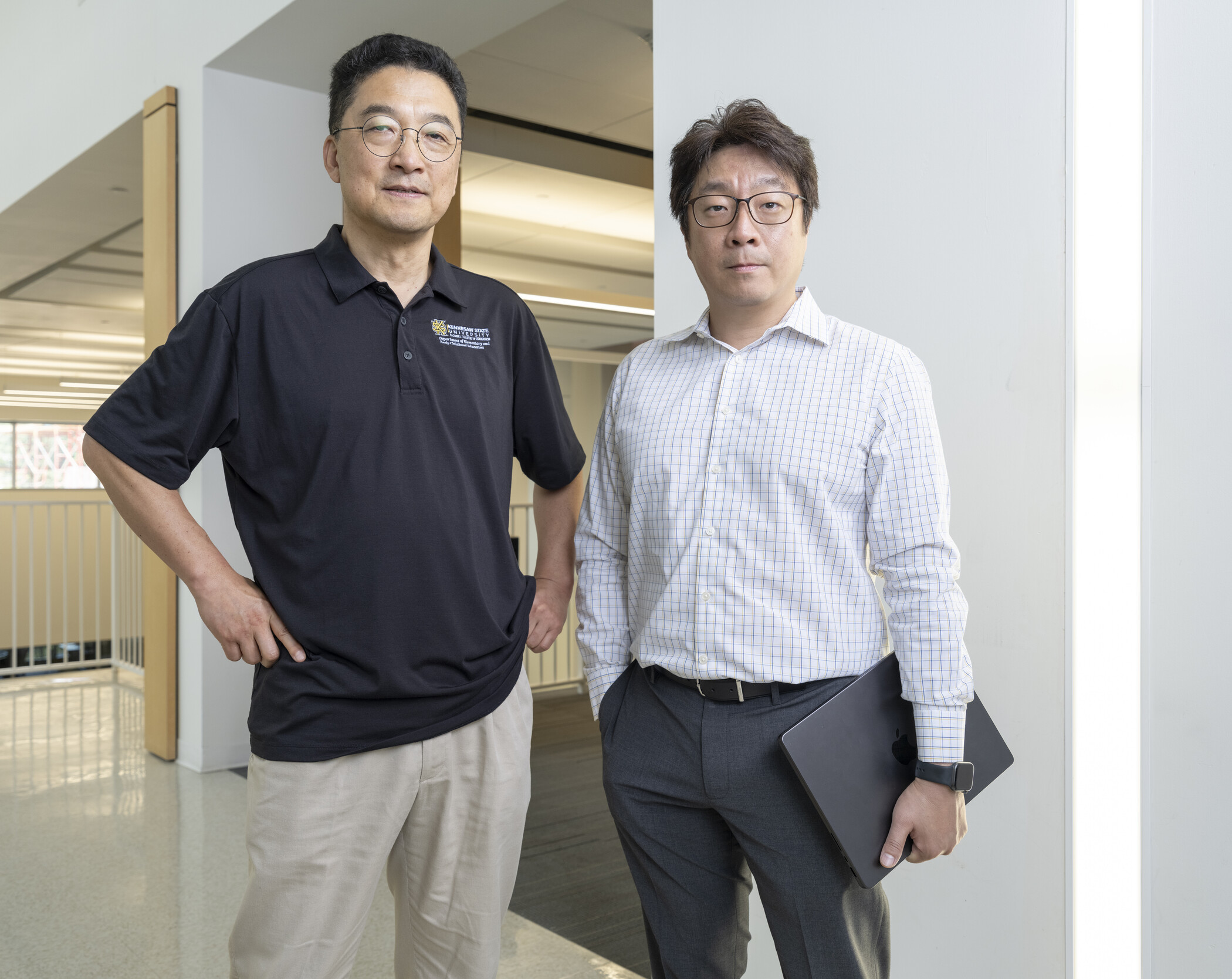 Two men stand indoors; one wears a black polo shirt and khakis, the other wears a white checkered shirt, gray pants, and holds a closed laptop. Both look at the camera in a well-lit hallway.