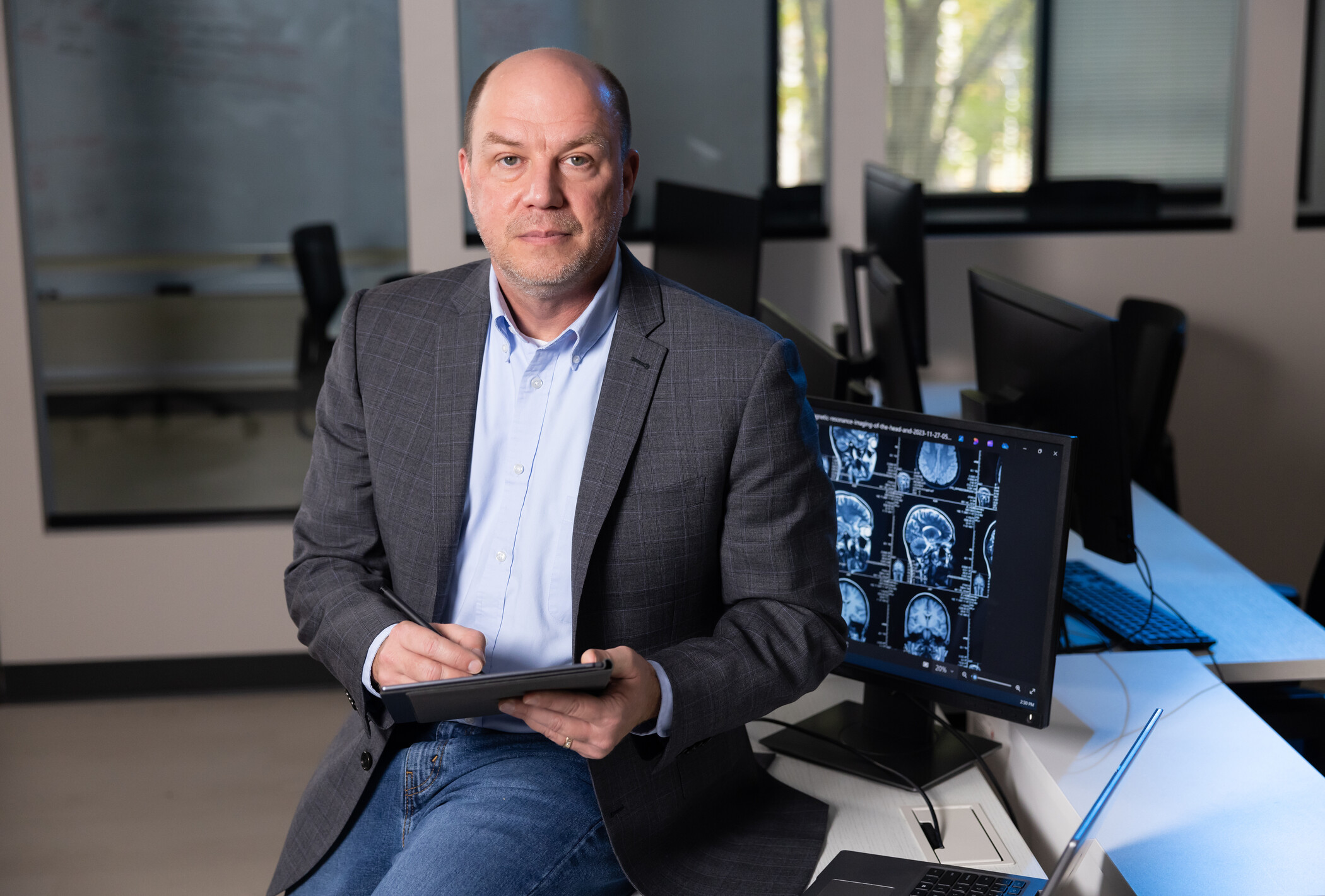 researcher sitting on table with tablet in hand. Background shows computer monitor with CT scans.