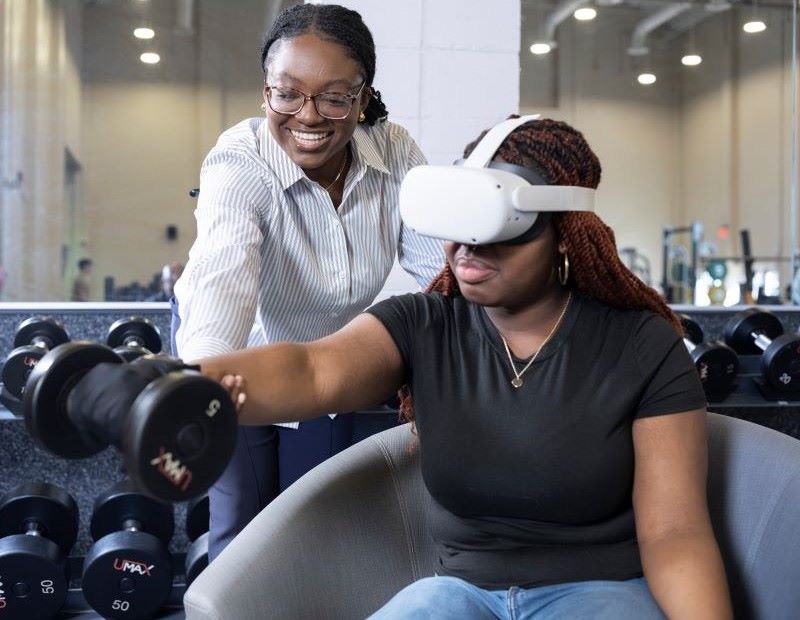 Person using VR headset lifts dumbbell with trainer nearby