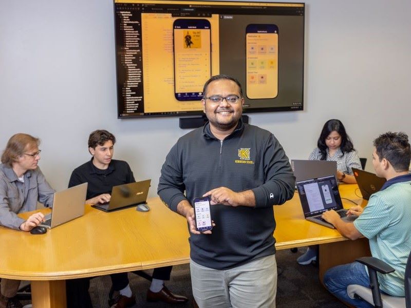A man stands in a conference room holding a smartphone displaying an app, while four others sit around a table working on laptops