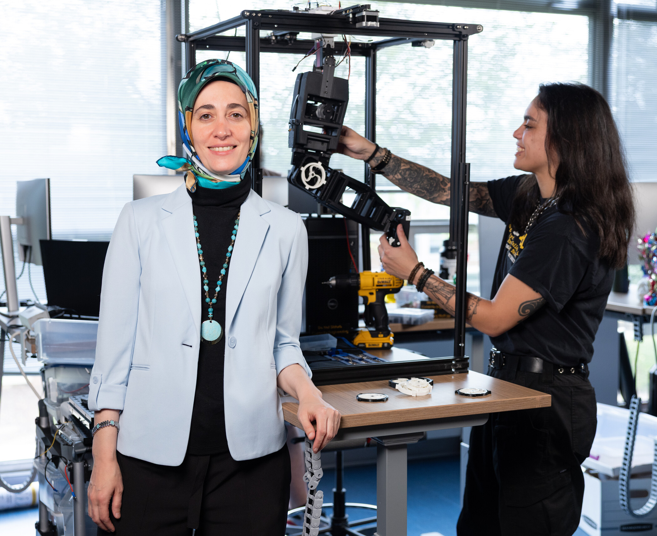 Two women in a lab: one stands in front smiling, the other adjusts a mechanical device on a table. Computers and technology equipment are visible in the background.