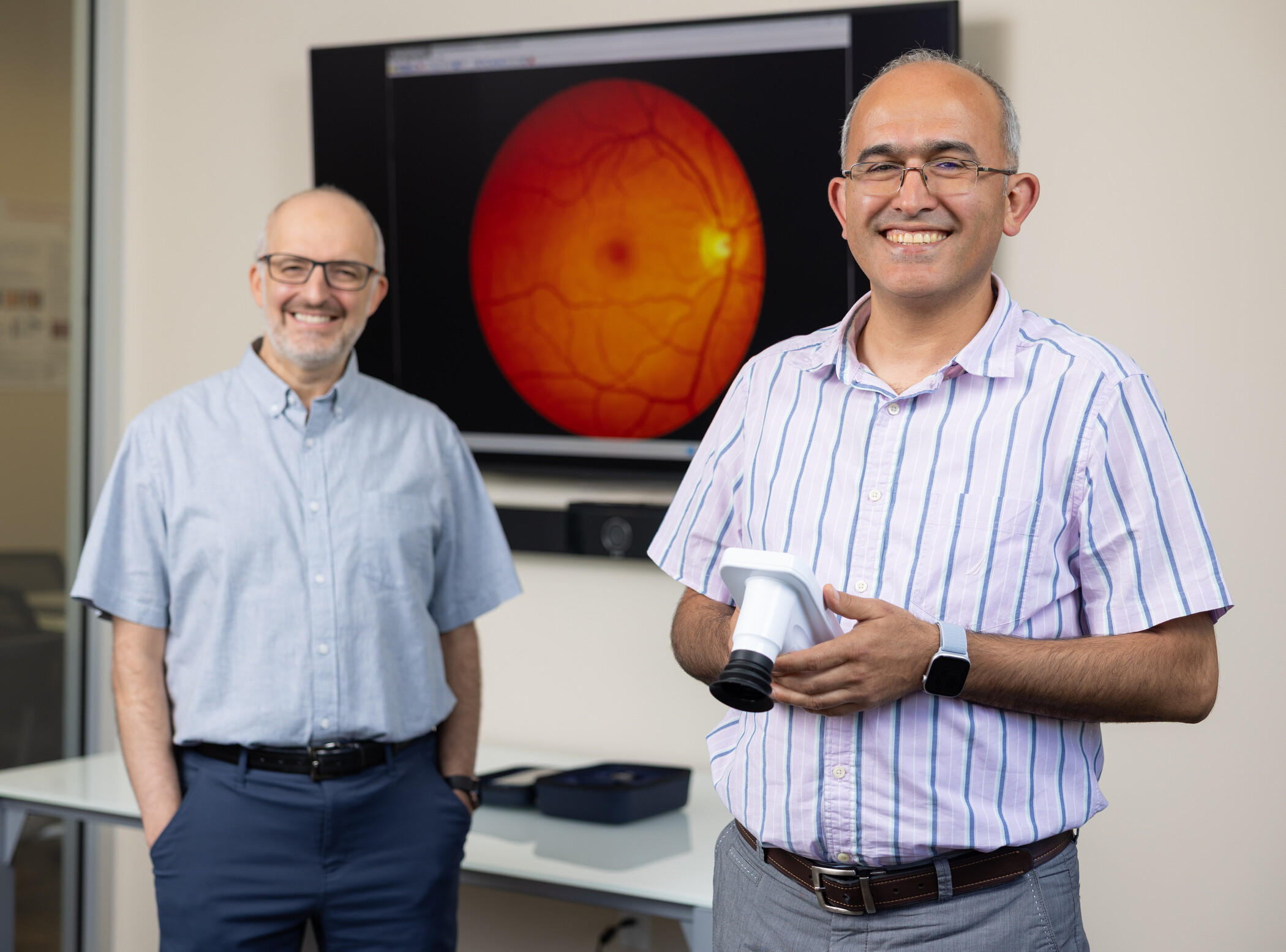 Two men stand in an office; one holds a medical device. A retinal image is displayed on a screen behind them.