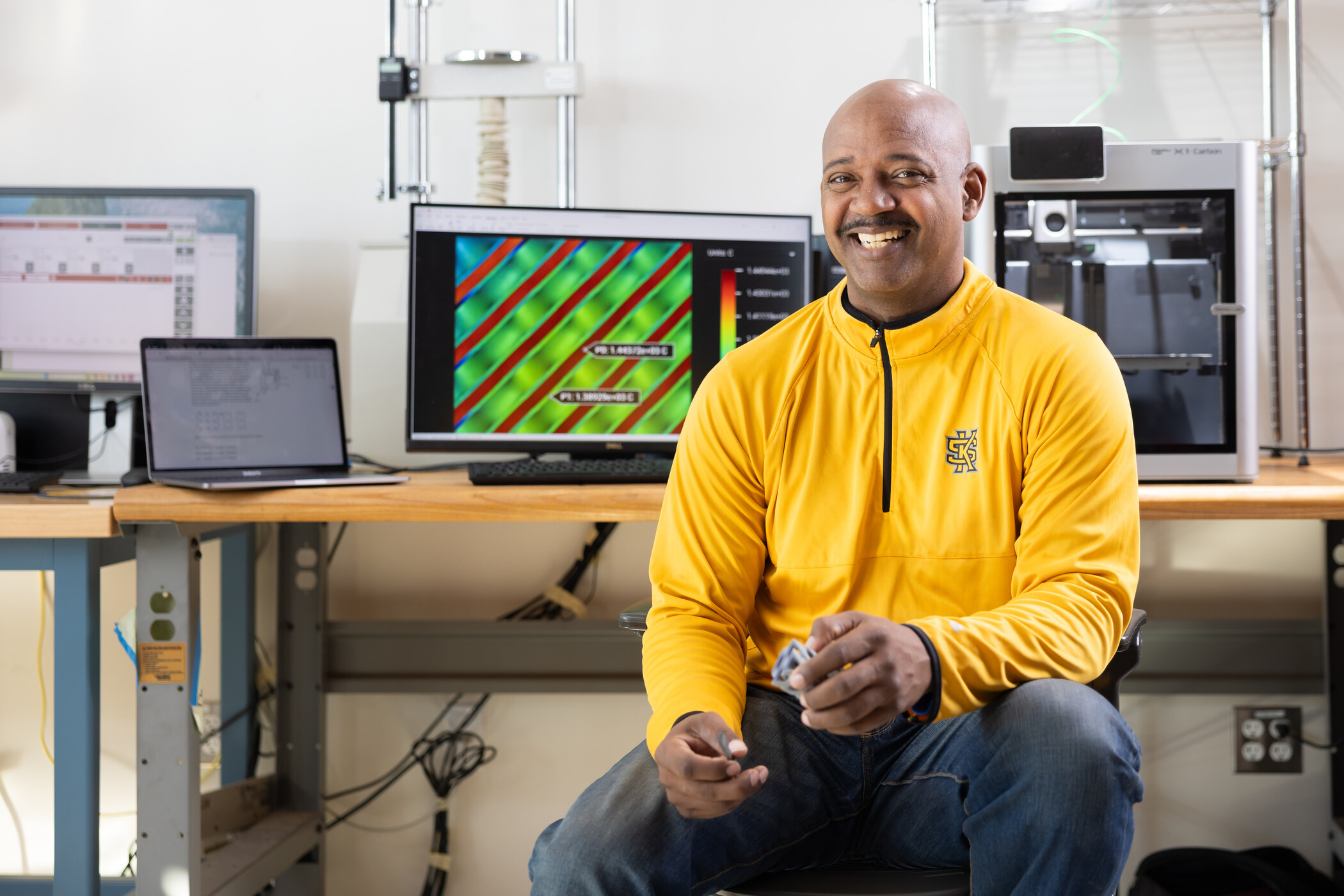 A smiling man in a yellow pullover sits at a workbench with two computer screens displaying data and a 3D printer in the background.