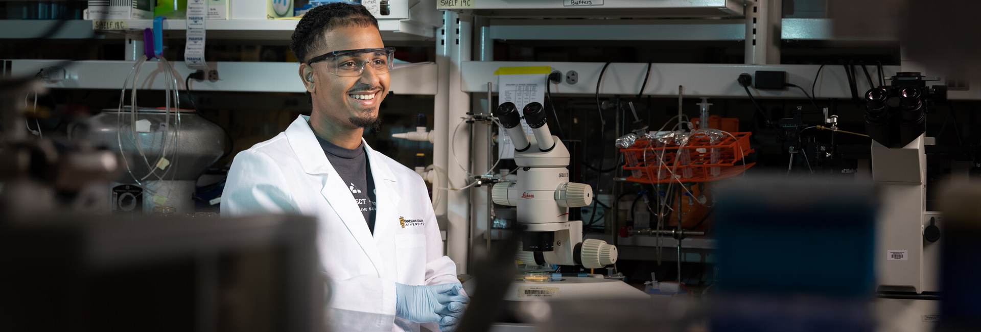 A student wearing a white lab coat and safety goggles smiles in a science lab, standing beside a microscope and other equipment.
