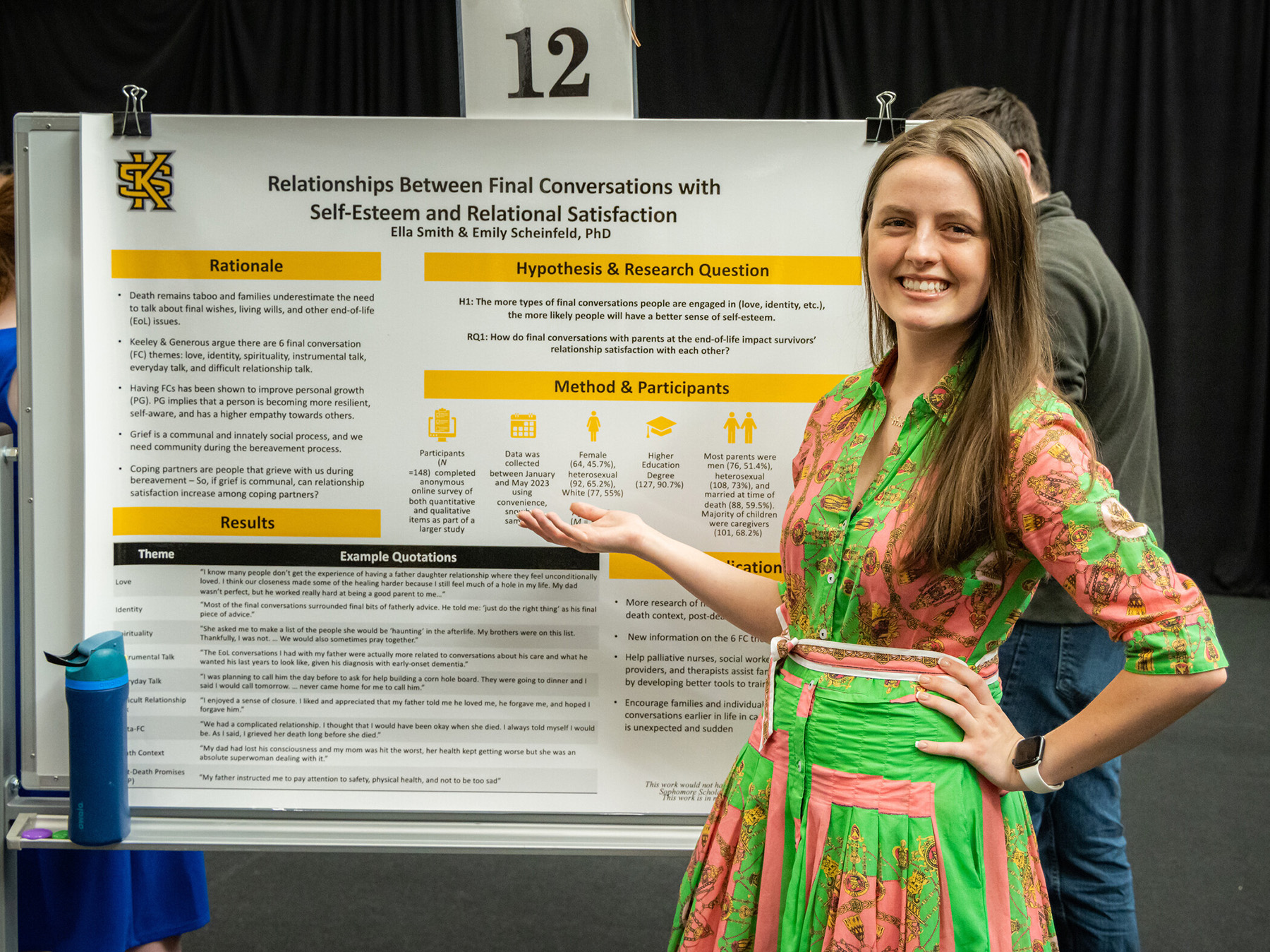 A student in a colorful dress stands beside a research poster titled "Relationships Between Final Conversations with Self-Esteem and Relational Satisfaction." She is smiling and gesturing towards the poster.