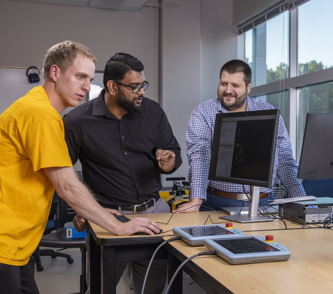 Three people collaborate in a lab environment, examining a computer screen. Two men, one in a yellow shirt and another in a black shirt, appear to be discussing while a man in a checkered shirt looks on.