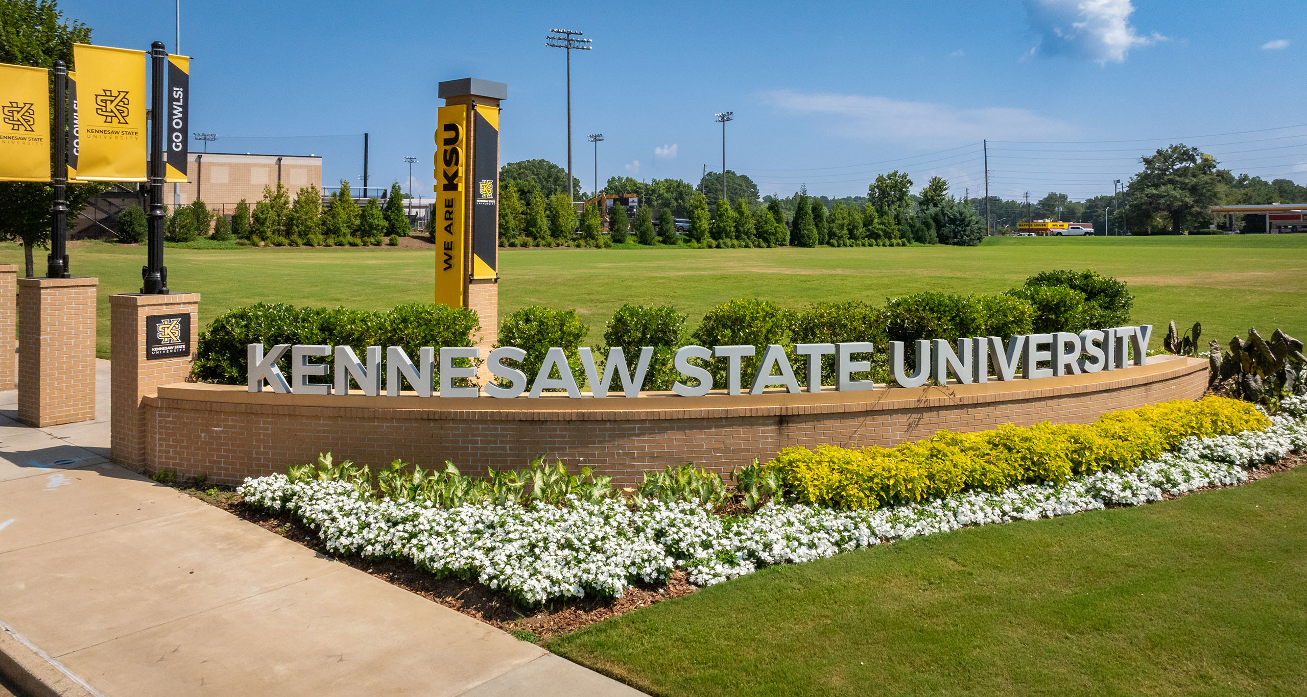 A sign for Kennesaw State University stands amid lush greenery and colorful flowers under a clear blue sky, exuding a welcoming and vibrant campus atmosphere.