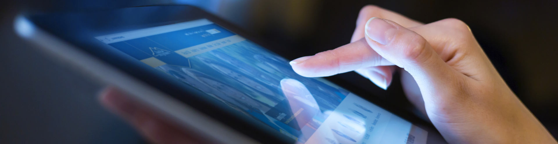 Close-up of a hand interacting with a glowing tablet screen in low light. The display shows charts, conveying technology and information access.