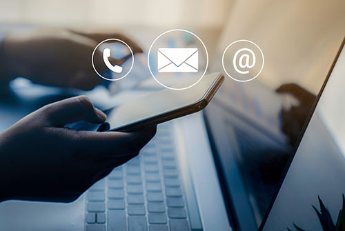 Hands holding a smartphone with contact icons for phone, email, and at-sign hovering above. A laptop is partially visible in the background.
